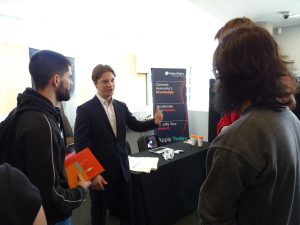 A picture of the Alphasights stand at last years careers fair. A man in a suit talks to two students 