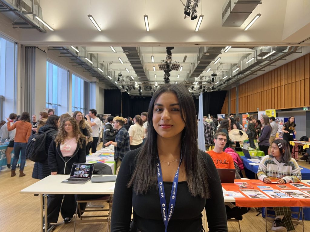 Archita Dwaram posing in front of the stalls at the Student Volunteering Fair. She is wearing a black top and a lanyard showing that she is a volunteer at the fair. 