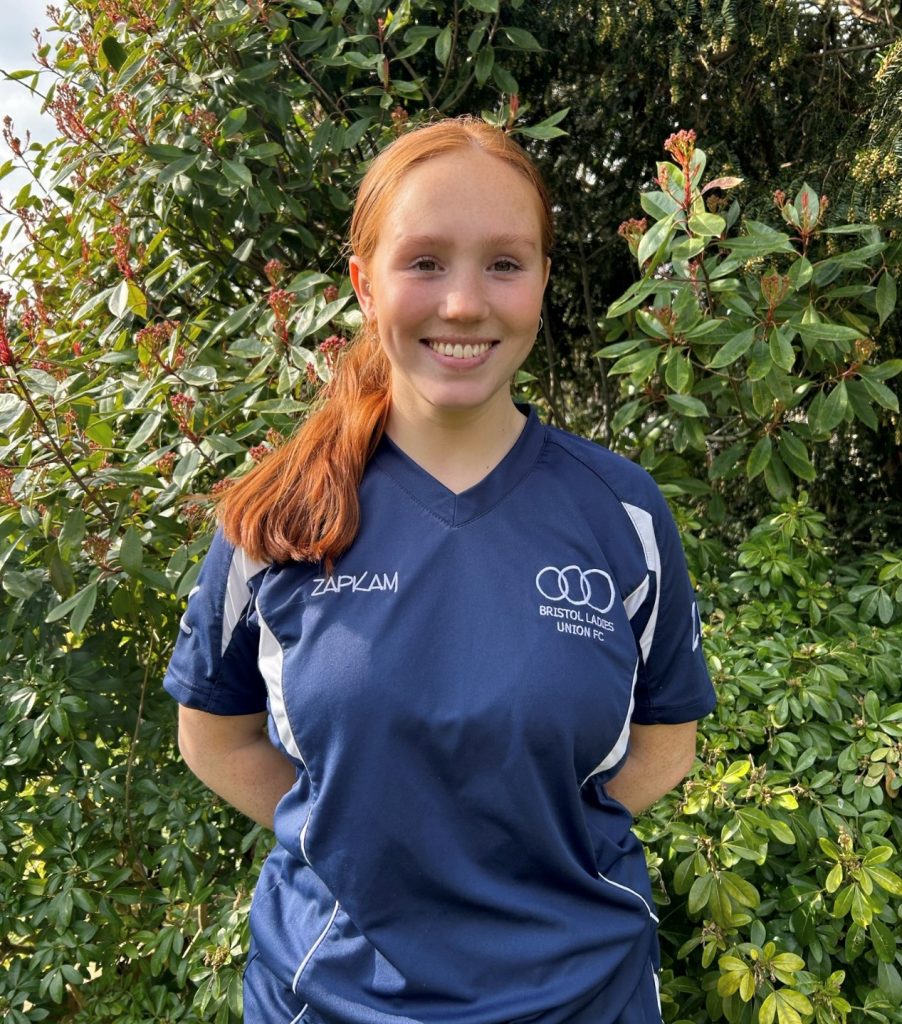 Ellie Haines standing in front of lush greenery. She has her hair in a ponytail and is wearing a blue and white football jersey.