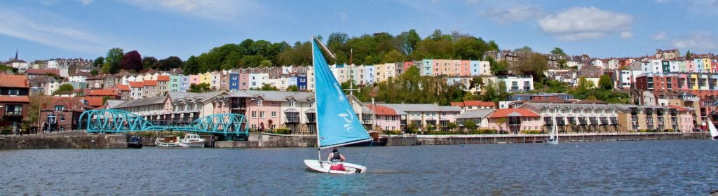 Bristol harbourside on a sunny day. There is a blue sailboat against a backdrop of colourful houses. 