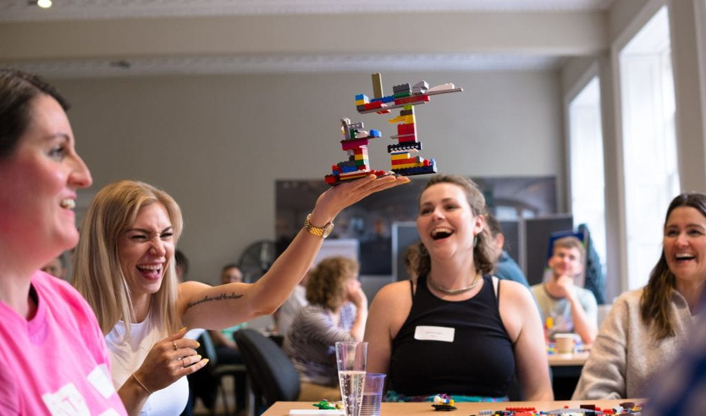 A photo of Izzie laughing in a group at a desk, as a colleague holds up a construction made of multi coloured plastic blocks.