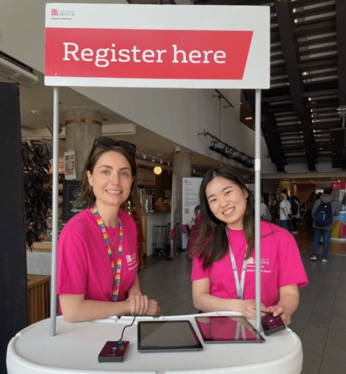 Two members of the Careers Service, both female, smiling underneath a signpost that reads "Register here"