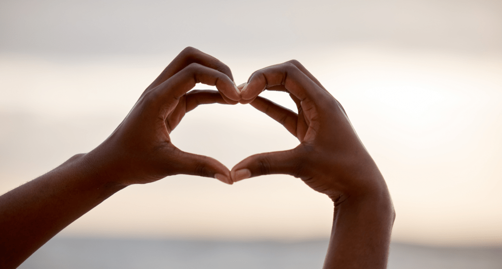Someone using their hands to make a heart shape, against a blurry background of the ocean and sky. 