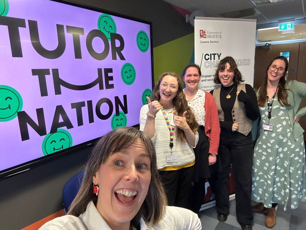 Five women look excited and happy standing by a large screen with the Tutor the Nation logo on.